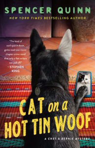 Cover of Cat on a Hot Tin Woof shows a black dog with a white ear holding his paw on a mobile phone that has the image of a cat on it.