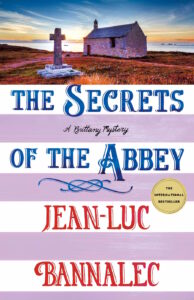 Cover of The Secrets of the Abbey shows a stone cross and stone chapel near a body of water.