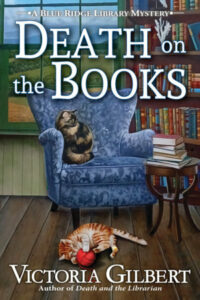 Cover of Death on the Books shows a cozy scene of a wing-back chair with a cat on it, a cat on the floor playing with a ball, a side table with a pile of books, and a bookshelf and window behind it. 
